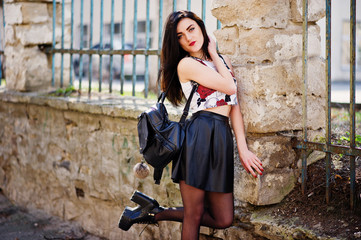 Young goth girl on black leather skirt and high heels punk shoes with backpack posed against iron fence.