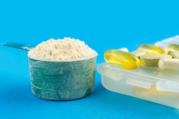 Protein powder in measuring plastic spoon and vitamin complex, omega 3, glucosamine capsules, supplements on blue background, sports nutrition
