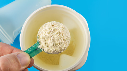 Male hand holds full measuring spoon with protein powder, directly above an almost empty container on blue background, sports nutrition