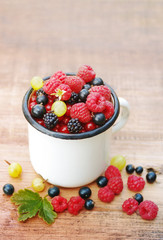 Fresh juicy berries, raspberries, currants, blackberries, a gooseberry in an old white iron mug on a wooden surface
