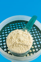 Protein powder in the measuring plastic spoon on top of the shaker bottle on blue background, sports nutrition