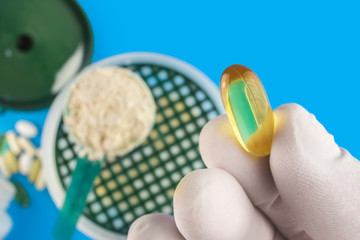 Hand in medical glove holds omega-3 capsule directly above a protein powder in the measuring spoon, shaker bottle with milk on blue background top view