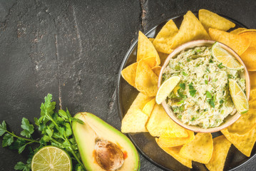 Homemade guacamole in bowl, served with tortilla nachos lime lemon and parsley, with half of avocado on black concrete table. Copy space