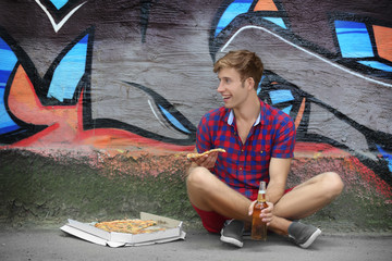 Young handsome man with pizza and beer sitting on asphalt on painted wall background