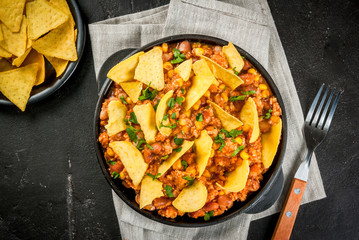 Traditional Mexican Chilean food, chili con carne, served in a portioned skillet pan with nachos tortilla. On black concrete table, copy space