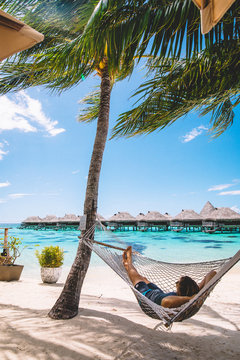 Man reclining in hammock by sea, distant beach huts, Mo'orea, South Pacific 