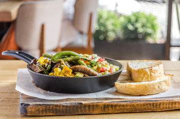 Omelet in a frying pan with vegetables, bread and cutlery on a wooden table, side view