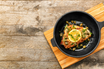 Frying pan with delicious chicken piccata on wooden board