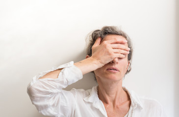 Middle aged woman with grey hair with hand covering face against white wall (selective focus)