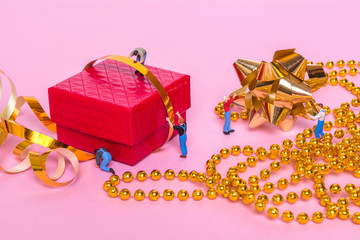 Miniature people and a gift box on a pink background. The process of packing gifts. Gold ribbon,...