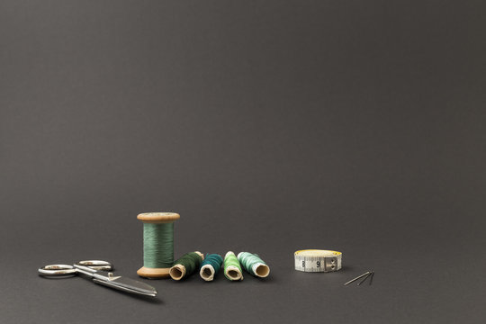 Different rolls with green polyester and silk thread, needles, scissors and a measuring tape with grey background