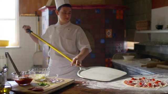 Slow motion shot of young chef in uniform carrying unbaked pizza with peel from table to wood fired oven in restaurant kitchen