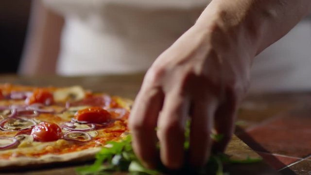 Tracking shot of male hand taking arugula leaves and scattering them on top of just baked pizza in slow motion