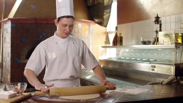 Tracking shot of young chef in uniform flattening dough with rolling pin on floury table in restaurant kitchen in slow motion