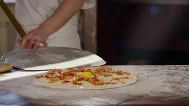 Tilt down slow motion shot of pizza maker putting dough base with toppings on peel from floury table in restaurant kitchen