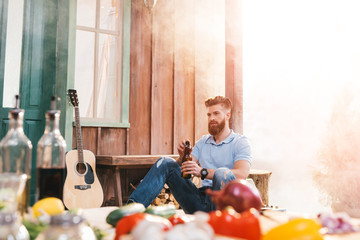 Handsome bearded young man sitting on porch and drinking beer