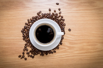 White cup with black, hot coffee standing on brown, roasted beans on wooden table