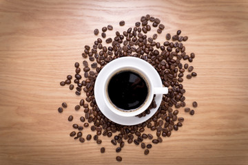 White cup with black, hot coffee standing on brown, roasted beans on wood table.