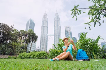 Rolgordijnen Reisconcept. Mooie jonge vrouw in hoed met smartphone zittend op het gras tegen Petrones Twins Tower in Kuala-Lumpur. © luengo_ua