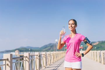 Healthy lifestyle and technology. Young pretty woman drinking water and listening music in earphones after jogging outdoors.