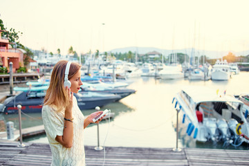 Fototapeta na wymiar Enjoying the sound. Happy young woman with earphones is listening music on broadwalk in marine harbor.