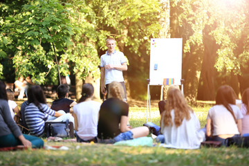 A group of young students in the park. View of a man gesticulating with his hands, standing against a defocused group of people sitting on the grass.