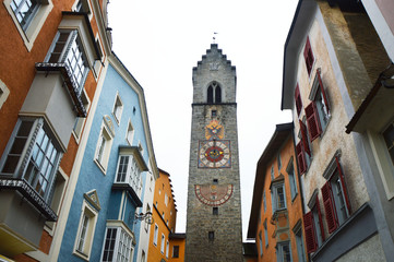 Fototapeta premium VIPITENO, ITALY - NOVEMBER 23, 2016: Medieval tower clock with colorful houses in Via Città Nuova, center of Vipiteno, historic village classified among the most beautiful in Italy, South Tyrol, Italy
