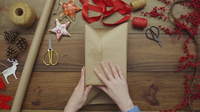 Aerial Lockdown Footage Of Woman Wrapping Christmas Present. Personal Perspective Of Female Preparing Gift For Occasion. Overhead Flat Lay Of Art And Craft Equipment Surrounding Lady's Hands.
