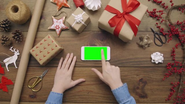 Aerial lockdown footage of woman using smart phone during Christmas. Personal perspective of female touching green screen on device. Overhead flat lay of gift wrapping equipment on wooden table.
