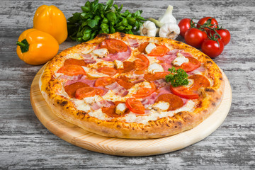 Pizza with sausage, chicken, bacon, ham, with rosemary and spices on a light wooden background. Italian pizza on a background of green basil and fresh vegetables