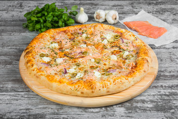 Pizza with salmon (red fish), with rosemary and spices on a light wooden background. Italian pizza on a background of green basil and fresh vegetables