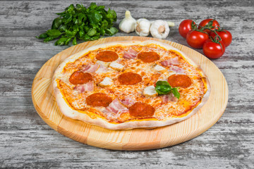 Thin pizza with salami (pepperoni), bacon and chicken with tomato sauce, rosemary and spices on a light wooden background. Italian pizza on a background of green basil and fresh vegetables