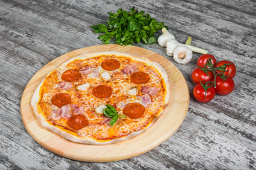 Thin pizza with salami (pepperoni), bacon and chicken with tomato sauce, rosemary and spices on a light wooden background. Italian pizza on a background of green basil and fresh vegetables