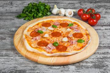 Thin pizza with salami (pepperoni), bacon and chicken with tomato sauce, rosemary and spices on a light wooden background. Italian pizza on a background of green basil and fresh vegetables
