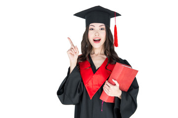 Beautiful young brunette woman in mortarboard holding book and pointing away with finger