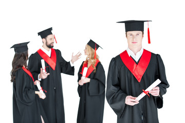 Young student in graduation cap with diploma and friends standing behind isolated on white