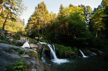 Fototapeta na wymiar Lovely wedding couple against waterfall on sunset at Carpathian mountains.