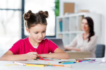 Mother looking at cute little girl drawing at table in office