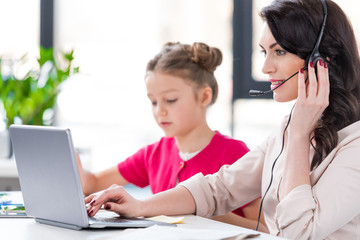 Cute little girl sitting near smiling mother in headset working with laptop