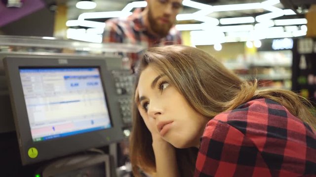 Stupid Displeased female cashier sitting by the cashbox in supermarket and to which the buyer is suitable