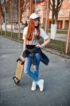 Young teenage urban girl with skateboard, wear on glasses, cap and ripped jeans at the yard sports ground.