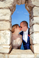Wedding. Happy couple day wedding. Beautiful bride and groom at the beach. Cheerful married couple standing and kissing on the stone wall window. Wedding couple staying over beautiful landscape.