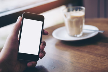Mockup image of businessman's hand holding black mobile phone with blank white screen on wooden table in vintage cafe while hot coffee latte cup on desk 
