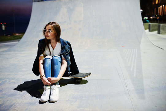 Young teenage urban girl with skateboard, wear on glasses, cap and ripped jeans at skate park on the evening.