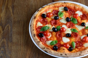 Top view of neapolitan pizza with pepperoni, mozzarella, cherry tomatoes and black olives on a wooden table for a dinner in italian restaurant. Close up. Macro. Photo can be used for menu design.