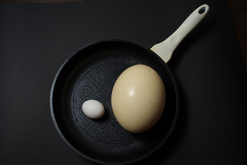 Ostrich and chicken eggs on a frying pan at black background