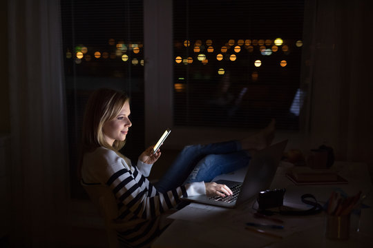 Woman At Desk, Holding Smartphone, Working On Laptop At Night.