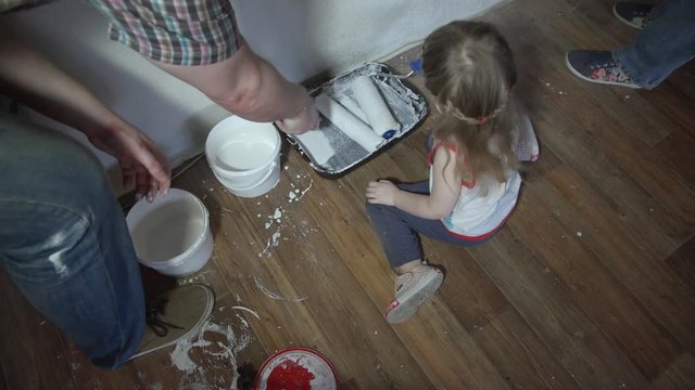 Little girl sitting on the floor next to the tray and buckets of paint, helps his parents done repair in the room. A young family paints a wall in their apartment, time lapse.