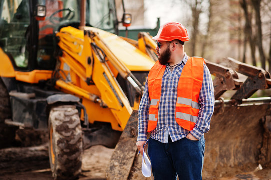 Brutal beard worker man suit construction worker in safety orange helmet, sunglasses against traktor with plan paper at hands.