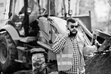 Fototapeta na wymiar Brutal beard worker man suit construction worker in safety orange helmet, sunglasses against traktor with mobile phone at hand.